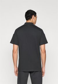 Μαύρο πόλο μπλουζάκι με κοντά μανίκια και απλό γιακά, κατασκευασμένο από απαλό ύφασμα, εμφανιζόμενο από πίσω, με ίσιο τελείωμα.