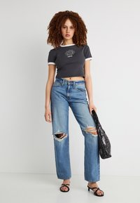 Νεαρή γυναίκα με κατσαρά μαλλιά, φοράει μαύρο κοντό T-shirt, σκισμένο μπλε τζιν, μαύρα σανδάλια και κρατάει μαύρη τσάντα χεριού.