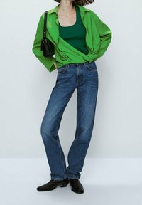 Chemise oversized verte superposée sur un débardeur vert, assortie à un jean en denim bleu et des bottines noires à cheville.