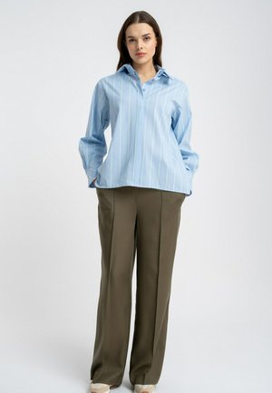 Młoda kobieta stoi, ubrana w jasnoniebieską koszulę w paski z kołnierzykiem oraz luźne, oliwkowozielone spodnie, na tle jednolitego tła.