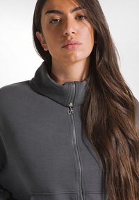 Giacca grigia con zip e colletto alto, realizzata in tessuto morbido e con una zip metallica dotata di un tirante circolare. Primo piano sul corpo superiore e sui capelli.