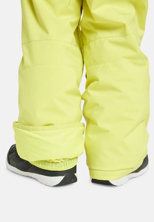 Gelbe wasserdichte Schneehosen mit umgeschlagenen Säumen und elastischen Knöcheln, kombiniert mit schwarzen Sportschuhen mit weißen Sohlen.