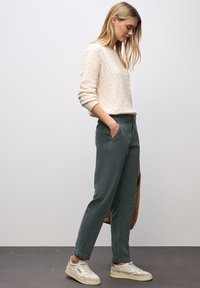 Kvinna med långt blont hår som bär en krämfärgad tröja, mörkgröna byxor och vita sneakers, stående med händerna i fickorna.