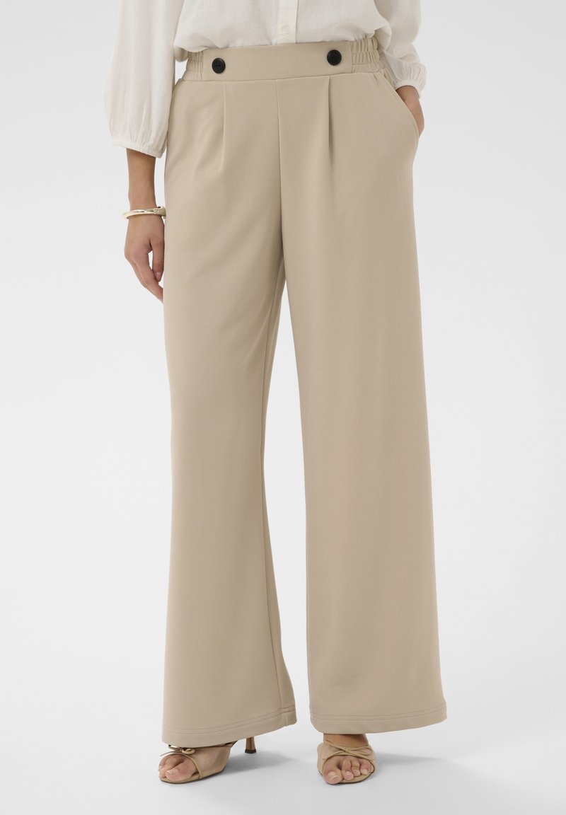 Personne portant un pantalon beige à jambes larges avec des boutons noirs à la taille, un chemisier blanc, un bracelet doré et des sandales à talons ouvertes beiges.