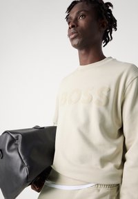 Sudadera beige con logo embosado "BOSS", puños acanalados y cuello redondo. Bolsa de cuero negro con asa en el fondo. Textura suave.