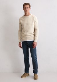 Cremefarbener, gerippter Pullover mit Rundhalsausschnitt, kombiniert mit blauen Jeans und beigen Slippern, auf einem neutralen Hintergrund stehend.