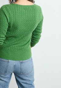 Maglione verde a trecce con orlo a coste, scollatura rotonda e maniche lunghe, abbinato a jeans azzurri a vita alta.