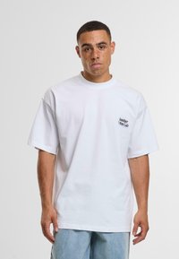 Camiseta blanca de manga corta hecha de algodón, con un pequeño logo negro en la parte izquierda del pecho y un diseño de corte relajado.