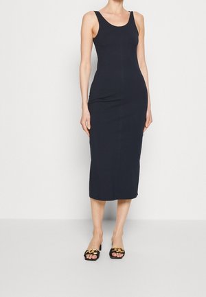 Marineblauwe mouwloze midi-jurk met een getailleerde pasvorm, gemaakt van een soepele stof. Gecombineerd met zwarte schoenen met gouden kettingaccenten.