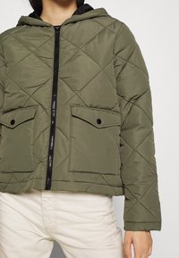 Oliwkowa pikowana kurtka puchowa z kapturem, z czarnym zamkiem błyskawicznym i dwoma klapowymi kieszeniami. Gładka tekstura z widocznymi przeszyciami.