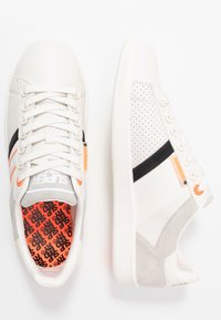 Zapatillas blancas con detalles en gris y negro, con laterales perforados, suelas blancas gruesas y una plantilla naranja brillante con patrón de logotipo negro.