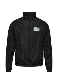 Veste noire zippée avec col montant, détails de couture rouges sur les manches, poignets et ourlet élastiques, et un patch rectangulaire blanc sur la poitrine.