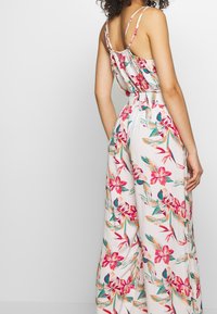 Combinaison florale avec des bretelles spaghetti fines, en tissu léger, taille élastique, et un motif tropical dans des nuances de rose, orange et vert.