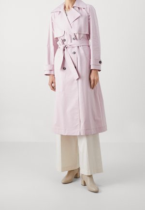 Vrouw draagt een lichtroze trenchcoat met riem en dubbele knopen, witte brede broekspijpen en beige enkellaarsjes, staand tegen een effen achtergrond.