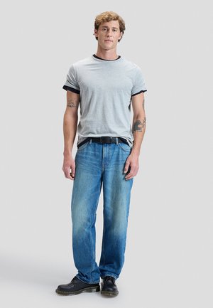 Junger Mann steht, trägt hellgraues T-Shirt, blaue Jeans, schwarzen Gürtel, schwarze Schuhe, mit Tätowierungen auf beiden Armen vor einfarbigem Hintergrund.