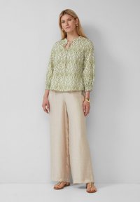 Kurzärmlige grüne Bluse mit weißem Blättermuster, rundem Ausschnitt und elastischen Bündchen, kombiniert mit beigen weiten Hosen und braunen Sandalen.