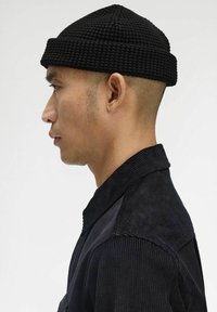 Uomo con berretto di tessuto nero e giacca di corduroy scura mostrato di profilo su uno sfondo neutro.