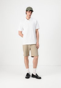 Weißes Polohemd mit kurzen Ärmeln, beiges Hemd mit umgeschlagenen Bündchen, schwarze Schuhe und eine olivgrüne Mütze. Das Model steht vor einem weißen Hintergrund.