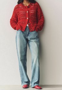 Vrouw in een rode gehaakte cardigan, lichtblauwe wijde jeans en rode schoenen, staand met handen in de zakken tegen een effen muur.