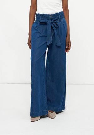 Γυναίκα που φοράει μπλε τζιν παντελόνι με ψηλή μέση και φαρδιά πόδια, με ζώνη δεμένη στη μέση, σε συνδυασμό με μπεζ παπούτσια με μυτερή μύτη και λευκό τοπ.