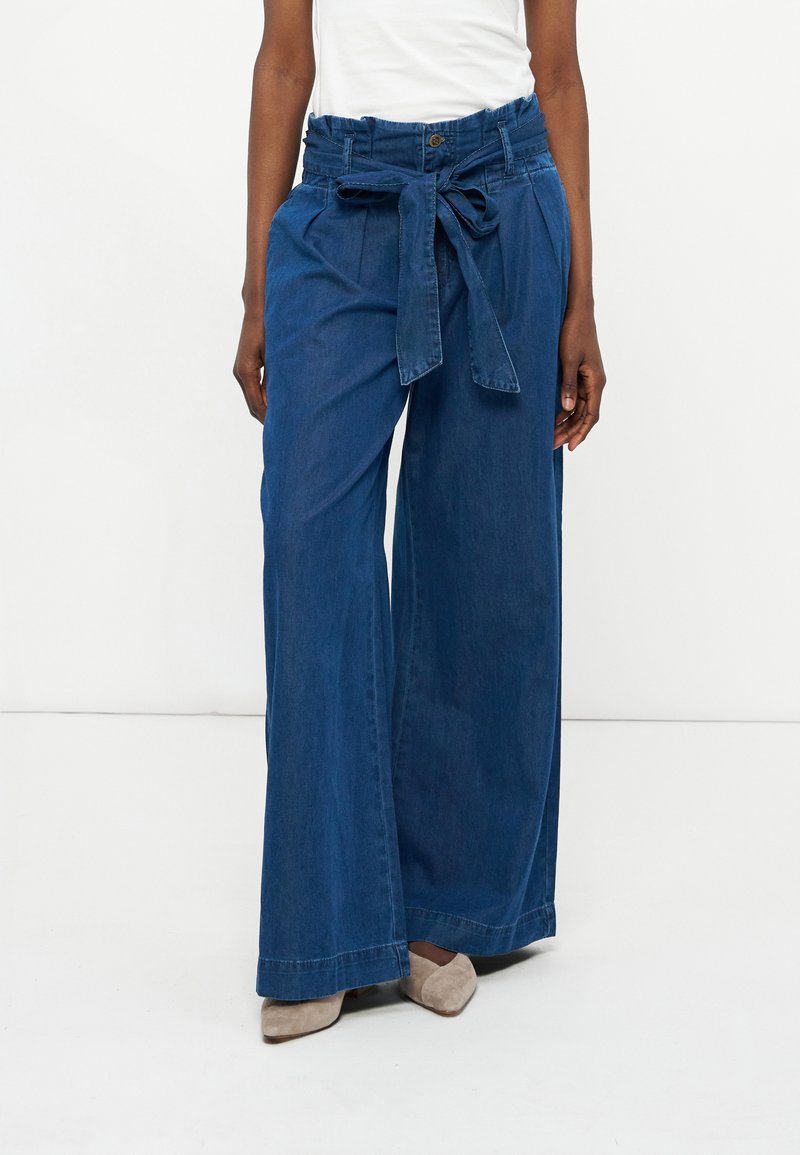 Γυναίκα που φοράει μπλε τζιν παντελόνι με ψηλή μέση και φαρδιά πόδια, με ζώνη δεμένη στη μέση, σε συνδυασμό με μπεζ παπούτσια με μυτερή μύτη και λευκό τοπ.