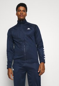 Tengeri kék tréningdzseki könnyű anyagból, elől cipzárral, magas gallérral és fehér logóval. Bordás mandzsetták és hem, kiegészítő csíkokkal.