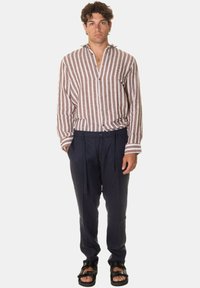 Camicia di lino a righe marrone e bianca con una vestibilità rilassata e una pattina; abbinata a pantaloni navy con coulisse e sandali neri.