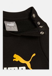 Musta puuvillainen T-paita, jossa on keltainen Puma-logo, napeilla varustettu kaulus sekä pehmeä pinta ja vahvistetut ompelut.