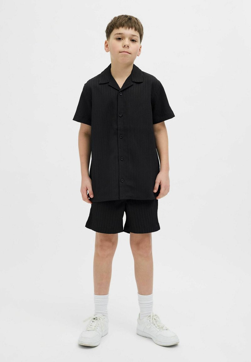 Camisa negra de manga corta con botones en el frente y rayas verticales, combinada con pantalones cortos a juego. Se lleva con zapatillas deportivas blancas y calcetines acanalados.