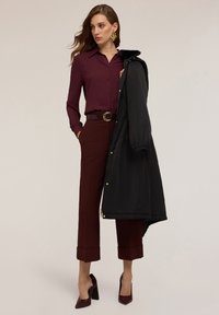 Blusa bordeaux con colletto, abbinata a pantaloni bordeaux su misura e un cappotto imbottito nero. Tacchi alti neri e grandi orecchini dorati completano il look.