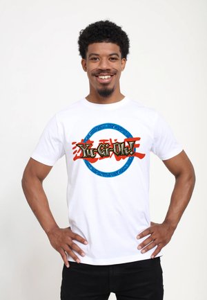 Mann trägt ein weißes T-Shirt mit einem bunten "Yu-Gi-Oh!"-Logo, lächelt mit den Händen in der Hüfte vor schlichtem Hintergrund.