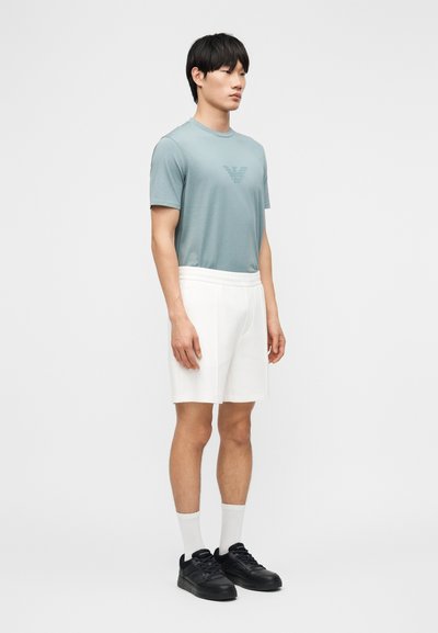 Άνδρας που στέκεται, φοράει ανοιχτό μπλε κοντομάνικο πουκάμισο με λογότυπο, λευκά σορτς, λευκές κάλτσες και μαύρα αθλητικά παπούτσια σε απλό λευκό φόντο.