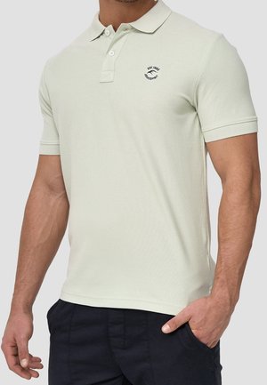 Mężczyzna w jasnobeżowej koszulce polo z krótkim rękawem, dwoma guzikami, logo na piersi oraz ciemnych spodniach, z ręką w kieszeni.