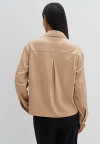 Beige suède shirt-jack met een relaxte pasvorm, overhemdkraag, rugplooidetail en lange mouwen, gecombineerd met zwarte broek.