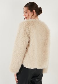 Beige nepbont jack met een zachte, fluffy textuur, ronde halslijn en lange mouwen, met een cropped silhouet.