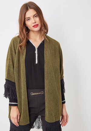 Kimono verde oliva de frente abierto con tejido texturizado y acentos de flecos negros en las mangas, combinado con una camiseta negra y pantalones de cintura alta.