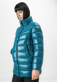 Vrouw in een teal gekleed gewatteerde jas met een hoge kraag, ritssluiting aan de voorkant en zijzakken, staand tegen een effen witte achtergrond.