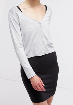 Kvinde iført en lysegrå cardigan med knapper over en sort tanktop, kombineret med en tætsiddende sort nederdel, stående mod en ensfarvet baggrund.