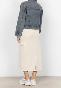 Donna che indossa una giacca di jeans grigia corta, una gonna midi beige con tasca laterale e sneakers bianche, in piedi su uno sfondo neutro.