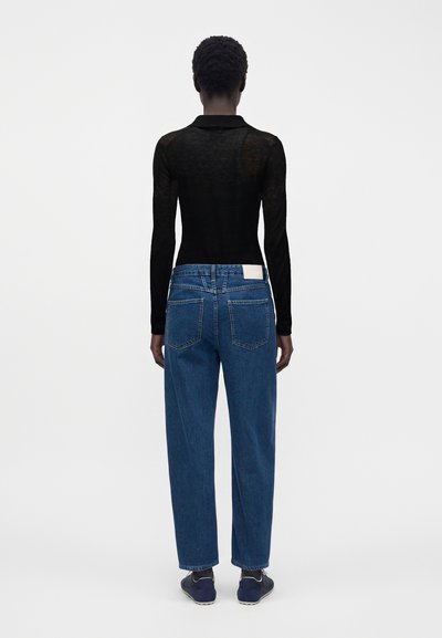 Personne de dos portant un haut noir transparent à manches longues, un jean bleu taille haute droit et des baskets foncées sur un fond clair uni.
