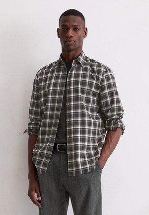 Chemise à carreaux dans des tons noirs, blancs et gris. Manches longues avec poignets retroussés, col à boutons, portée par-dessus une chemise noire. Coupe ajustée.