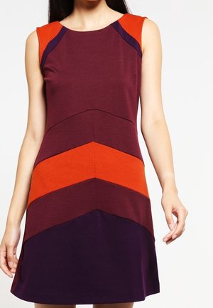 Robe sans manches avec motif chevron géométrique en violet foncé, bordeaux et orange vif, portée par une personne debout.