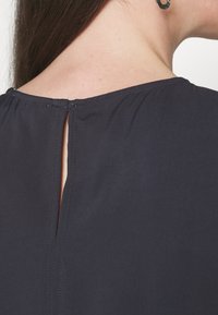 Gros plan de l'encolure dos sur un vêtement noir avec une petite fente verticale et une boucle d’oreille créole noire visible sur une personne aux cheveux bruns.
