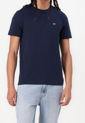 Mand iført mørkeblå t-shirt med korte ærmer og lille hvid "GAP"-logo på brystet samt lyseblå jeans mod hvid baggrund.