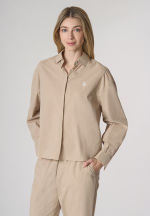 Camicia beige con bottoni, colletto e maniche lunghe. Realizzata in tessuto leggero, presenta un logo ricamato sul petto.