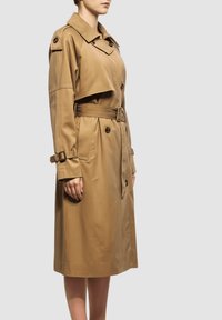 Mujer que lleva un trench beige de longitud a la rodilla con una cintura ceñida por un cinturón, frente abotonado y detalles de correa con hebilla en las mangas y los hombros.