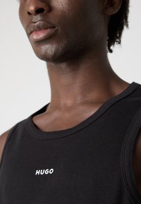 Tricou fără mâneci, din material texturat negru, cu un logo alb "HUGO" pe piept, guler rotund și o croială ajustată pe umeri și gât.