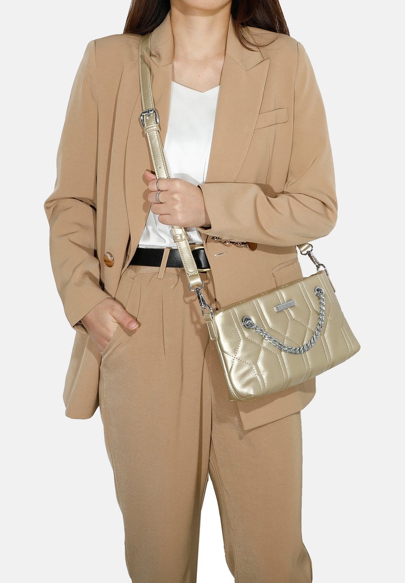 Borsa in metallo dorato con design trapuntato, caratterizzata da una tracolla e accenti in argento, tenuta accanto a un blazer beige e pantaloni.