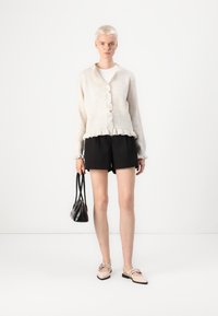 Cardigan bej tricotat, cu închidere cu butoane și tiv volanat, asortat cu pantaloni scurți negri și pantofi slip-on bej. Ține o geantă mică neagră.