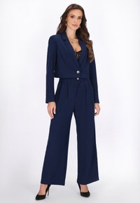 Woman with long wavy hair wearing a navy cropped blazer and high-waisted wide-leg trousers, black lace top underneath, and black pointed heels.